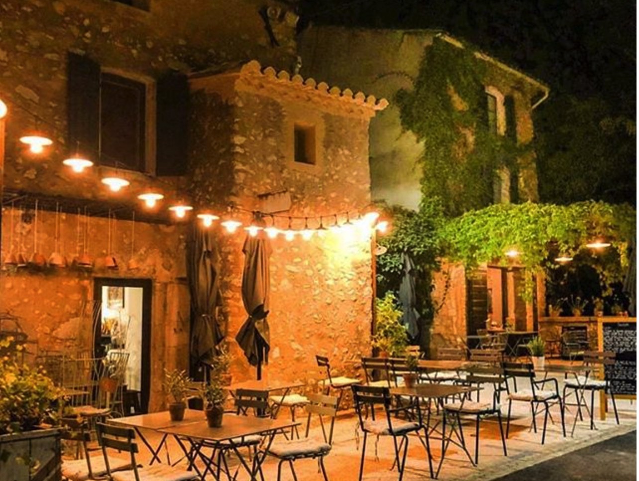 An enchanting outdoor dining area at night, illuminated by string lights, featuring empty tables and chairs set under a vine-covered building facade.